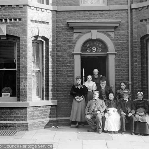 Family group posing outside of house, above door sign reads 'M. Eden, 9'
