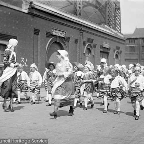 Women and children in costume in street procession outside building with sign for 'Tramway garage.'