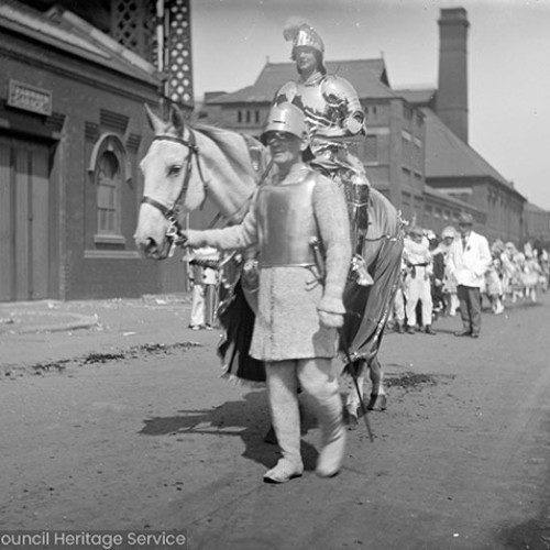 Two men dressed as knights, one on horse, outside building with sign for 'Tramway garage.'