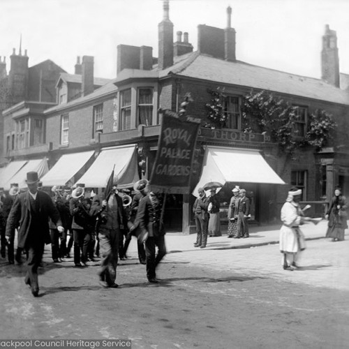 Crowds in street scene, including a placard for 'Royal Palace Gardens.'