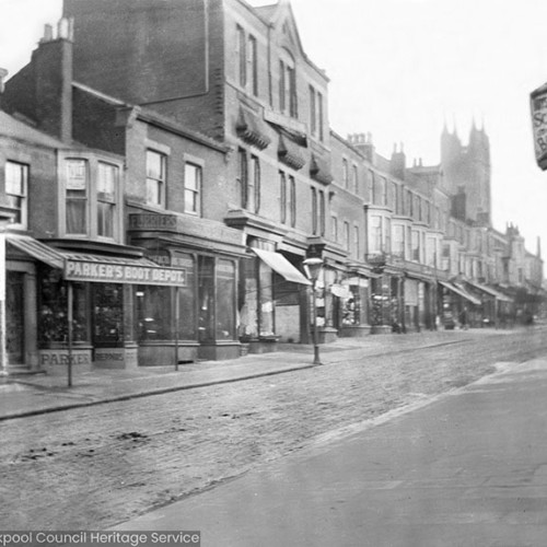 Street scene with shops including 'Parkers Boot Depot' and a sign for 'Boys and Girls School Boots.'