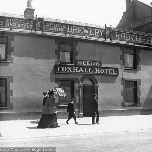 People walking in front of hotel building with signs reading 'Seeds Foxhall Hotel' and 'Fine Ales and Stout, Spring Lane Brewery, Radcliffe.'