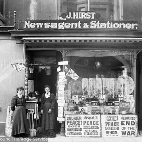 Two women standing outside of 'J. Hirst, Newsagent & Stationer.' Newspaper Headlines read 'Courier: Peace! Boer Surrender, Universal Rejoicing', 'Dispatch: Peace Declared, Official Dispatches', 'Echo: Peace Arranged, Official', Chronicle: End of the War'.