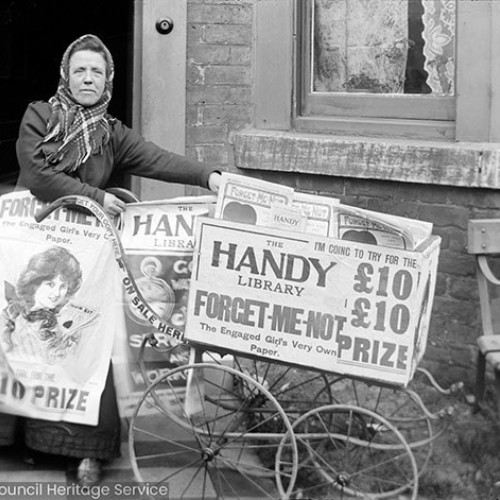 Woman with hand cart selling newspapers, advertisement on cart reads 'The Handy Library. Forget-Me-Not, the Engaged Girls very own paper. I'm coming to try for the £10 Prize.'