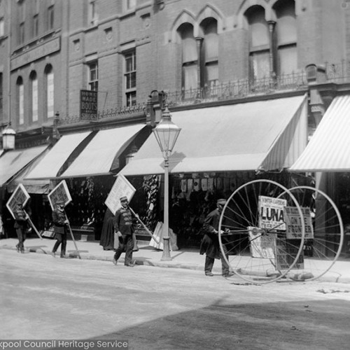 Men carrying placards in street, signs read 'Luna' and 'On the Frontier.' Shop advert in background reads 'Home made boots and shoes.'