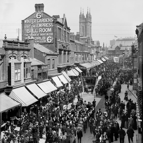 protest march being watched by crowds in street, advertisement on wall reads '6D. Winter Gardens, Visit the Empress Ballroom, the Grandest Sight in the World. Admission 6D.'