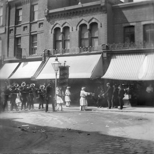 Street scene with parade including placard reading 'Royal Palace Gardens.'