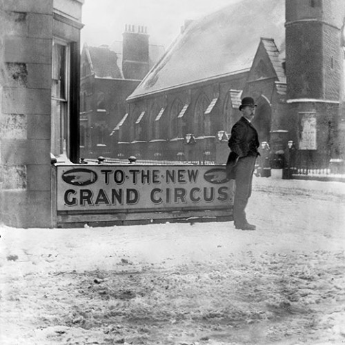 Man in a street standing in front of advert 'To the New Grand Circus'.