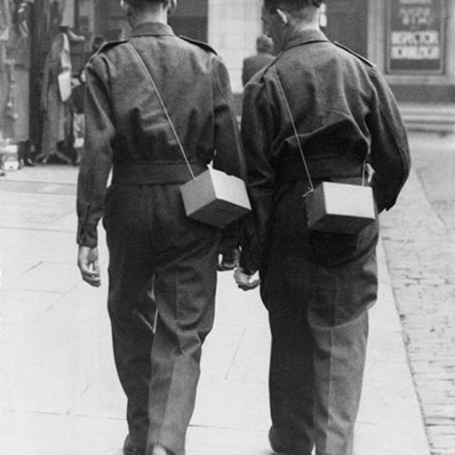 Two soldiers walking up the street wearing uniform and each carrying a gas mask box over their shoulder.