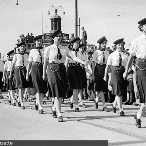 Group of girls in uniform, marching past onlookers.