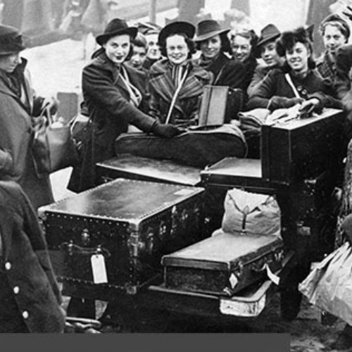 Group stood surrounding their luggage which is on a trolley being pulled by a railway worker.