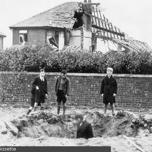 Child stood in a crater caused by bomb damage, other children stood watching. Behind them is a wall and a house which has almost been completely destroyed.