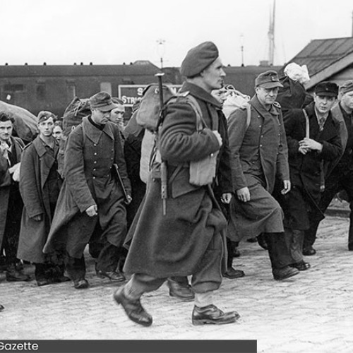 Group of men walking up a hill carrying bags on their backs. The man at the front of the group is carrying a rifle. A train can be seen in the background.