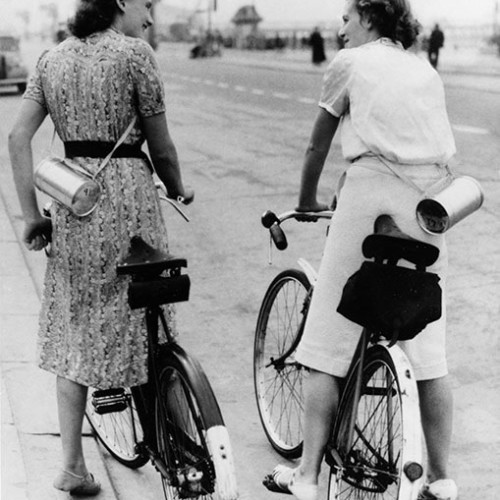 Two women on bicycles stopped on the Promenade and carrying a gasmask box over their shoulder.