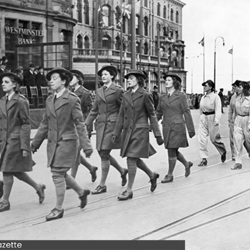 Group of women marching in formation past the Town Hall. The group is split into two, with the front group wearing a different uniform to the ones behind them.