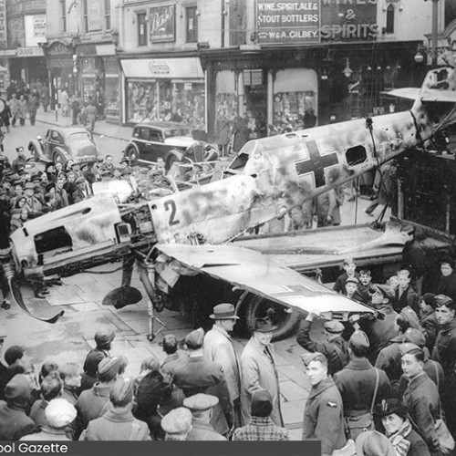 Damaged aeroplane on the street, next to shops surrounded by a crowd of onlookers.