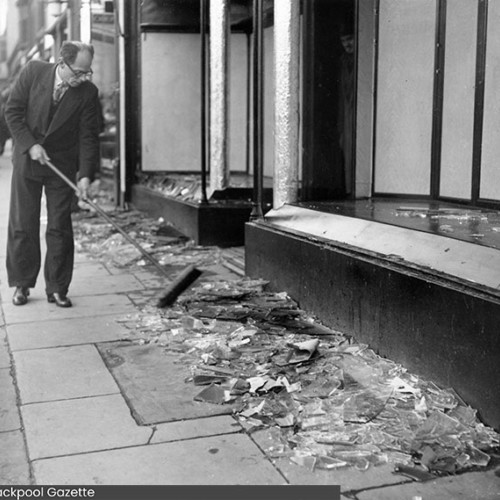 Man sweeping up glass on the pavement outside of a shop.