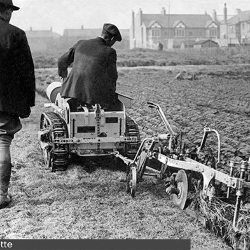 Man sat on a small tractor which is pulling a plough across a field. Another man is walking beside the tractor.