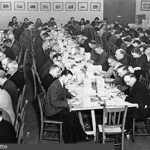 Room filled with rows of tables and workers with stacks of paper in front of them.