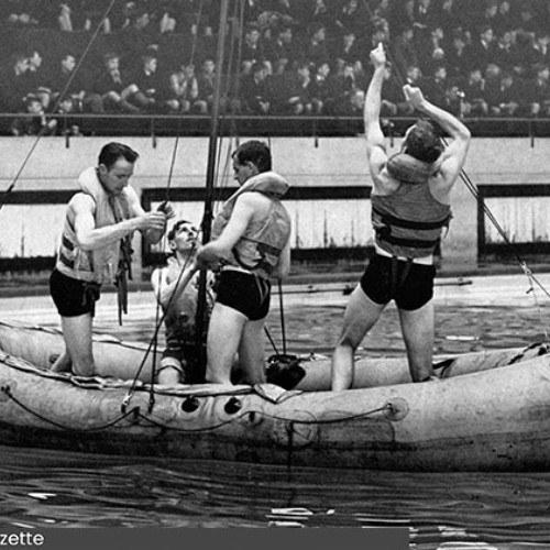 Swimming pool with spectators sat in the stands watching a group of four men who are in an inflatable boat in the swimming pool.