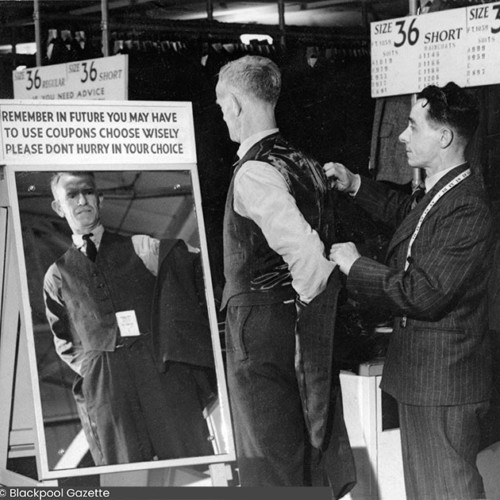 Tailor with a tape measure around his neck, putting a suit jacket on to a man who is looking into a mirror. Above the mirror reads, Remember in future you may have to use coupons choose wisely please don't hurry in your choice.