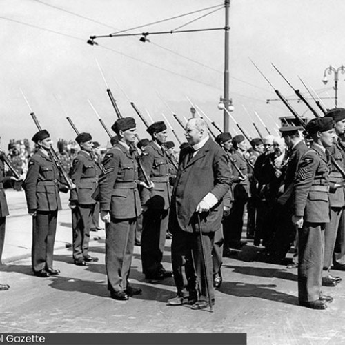 Man in suit with a walking stick inspecting a group of men in uniform with rifles on their shoulders.
