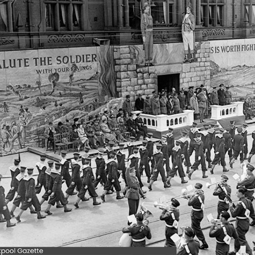 Entrance to the Town Hall which has been covered in painted boards which read Salute The Soldier With Your Savings. Spectators are watching a march past and a military band playing instruments.