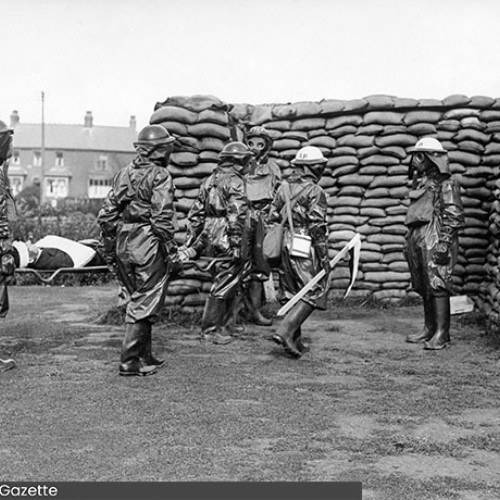 Group of wardens wearing gas masks and other protective clothing, stood in front of a sandbag wall.
