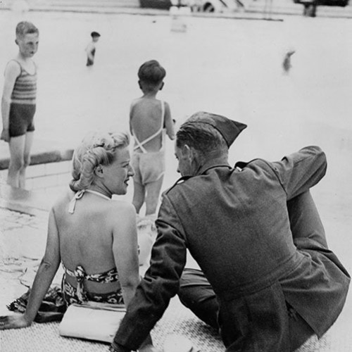 Solider sat talking to a lady in a swimsuit on the edge of a swimming pool, with children playing in the water behind them.