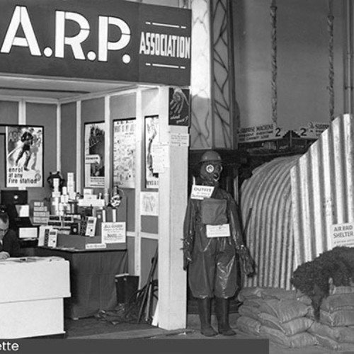 Man sat at a table in a stall with a large sign above it saying Blackpool A.R.P Association. To the right is an entrance to an air raid shelter with sandbags in front.