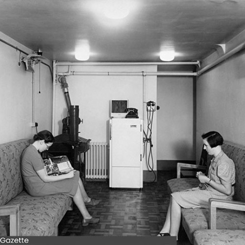 Two ladies sat opposite each other on benches in an air raid shelter. The lady on the left hand bench is reading a magazine, whilst the lady on the right hand bench is knitting.