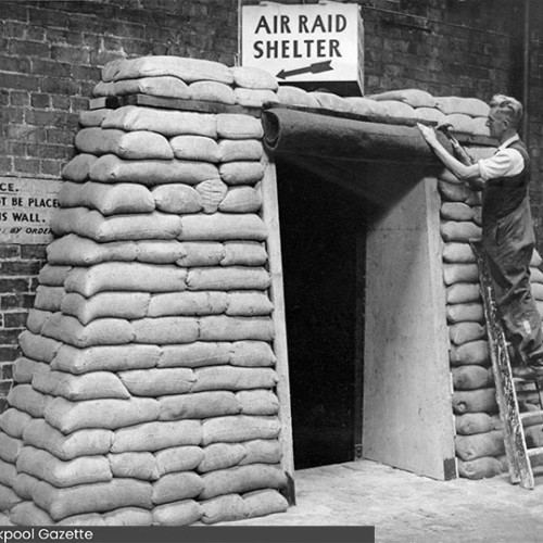 Sandbagged entrance to an air raid shelter, with a man up a ladder using a hammer to fix something above the doorway.