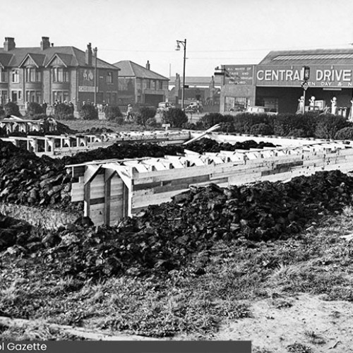 Field with large air raid shelters constructed on it, with houses and the Central Drive Garage behind the field.