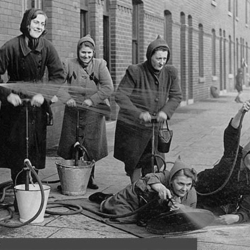 Group of seven women practicing fire drills. One woman who is wearing a Wardens helmet is guiding the group, whilst three of the women are pumping water and the other three are pointing the hoses with one of them stood and the other two laying on the floor.