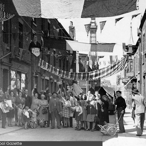 Street decorated with flags and people stood in the street celebrating.