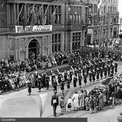 Group of sailors on parade outside the Town Hall which has a banner saying Welcome HMS Penelope. A large crowd of onlookers surround the sailors.