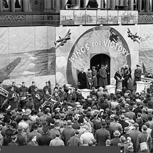 Large crowd, military band and dignitaries stood outside the Town Hall entrance. The entrance has been covered with boards that have been painted with the words Wings for Victory.