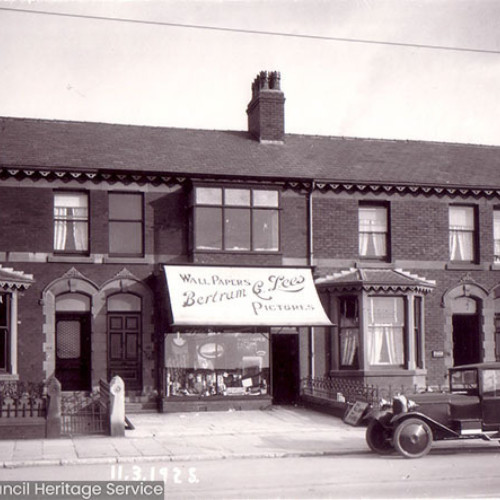 Street scene of a row of houses with a shop in the centre with a vintage car parked outside.