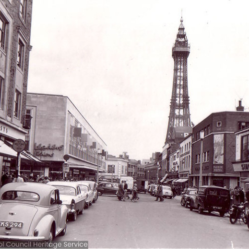 Street scene busy with people and parked cars, with Blackpool Tower in background.