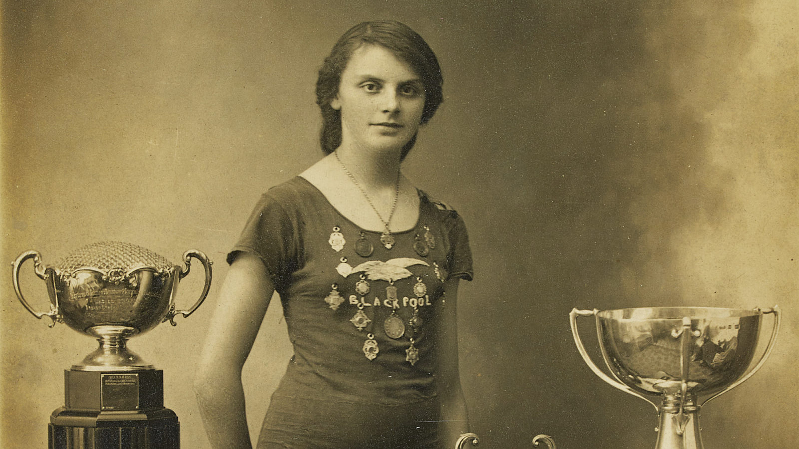 Black and white photograph of Lucy Morton stood wearing her swimming costume, which has the word Blackpool and multiple medals sewn on the front. She is stood between a stand with a trophy on and a table with two trophies and a number of medals on. The photograph has been signed Yours Sincerely Lucy 1914.