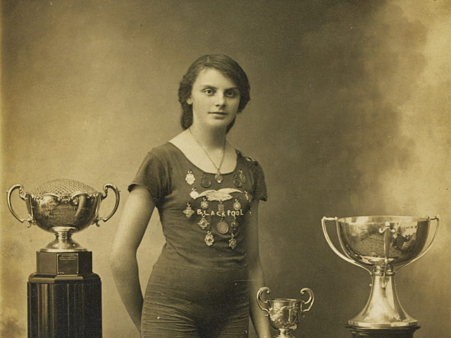 Black and white photograph of Lucy Morton stood wearing her swimming costume, which has the word Blackpool and multiple medals sewn on the front. She is stood between a stand with a trophy on and a table with two trophies and a number of medals on. The photograph has been signed Yours Sincerely Lucy 1914.