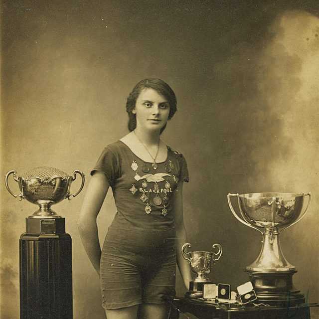 Black and white photograph of Lucy Morton stood wearing her swimming costume, which has the word Blackpool and multiple medals sewn on the front. She is stood between a stand with a trophy on and a table with two trophies and a number of medals on. The photograph has been signed Yours Sincerely Lucy 1914.