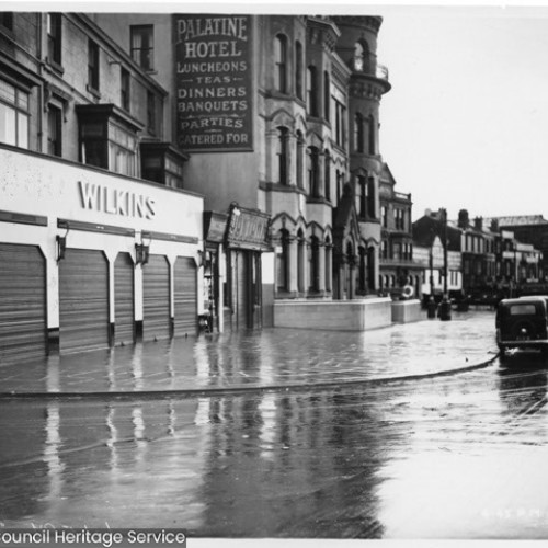 The Wilkins shop front with closed shutters and the Palatine Hotel, which has a large advertisement of their services on their side wall.