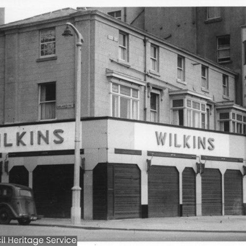 Wilkins shop front with closed shutters and a car parked outside. To the right of Wilkins is the Old England Cafe Snack Bar and the side wall of the Palatine Hotel which has a large advertisement on listing the hotel facilities.