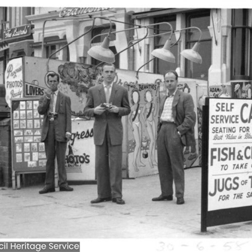 Three men, two of which have cameras are stood by their photography business and next to a sign advertising the Cafe next door.