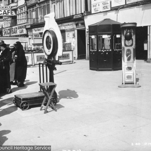 Machines and a group of people on the forecourt in front of a row of buildings.