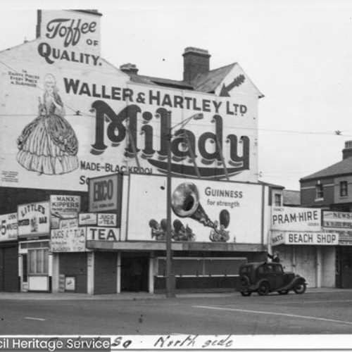 Multiple shop fronts surrounding street corner, with a large advertisement for Toffee of Quality Waller & Hartley Ltd Milady filling the side wall of a building.