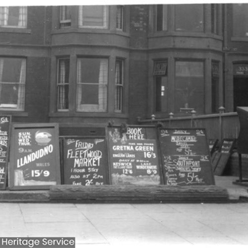 Advertising boards for numerous coach trips lined up outside of a row of buildings.