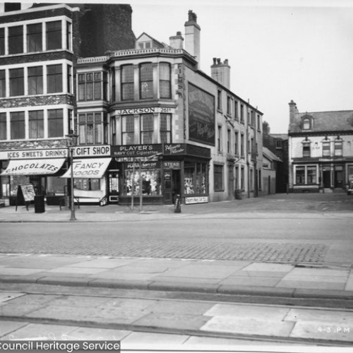 Street corner which leads into a square area. Shop fronts to the left advertise Ices, Sweets, Drinks, Chocolate, Gift Shop, Fancy Goods.