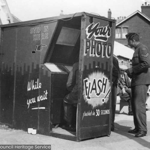 Man in military uniform stood outside a photo booth, whilst another man is sat inside the booth.